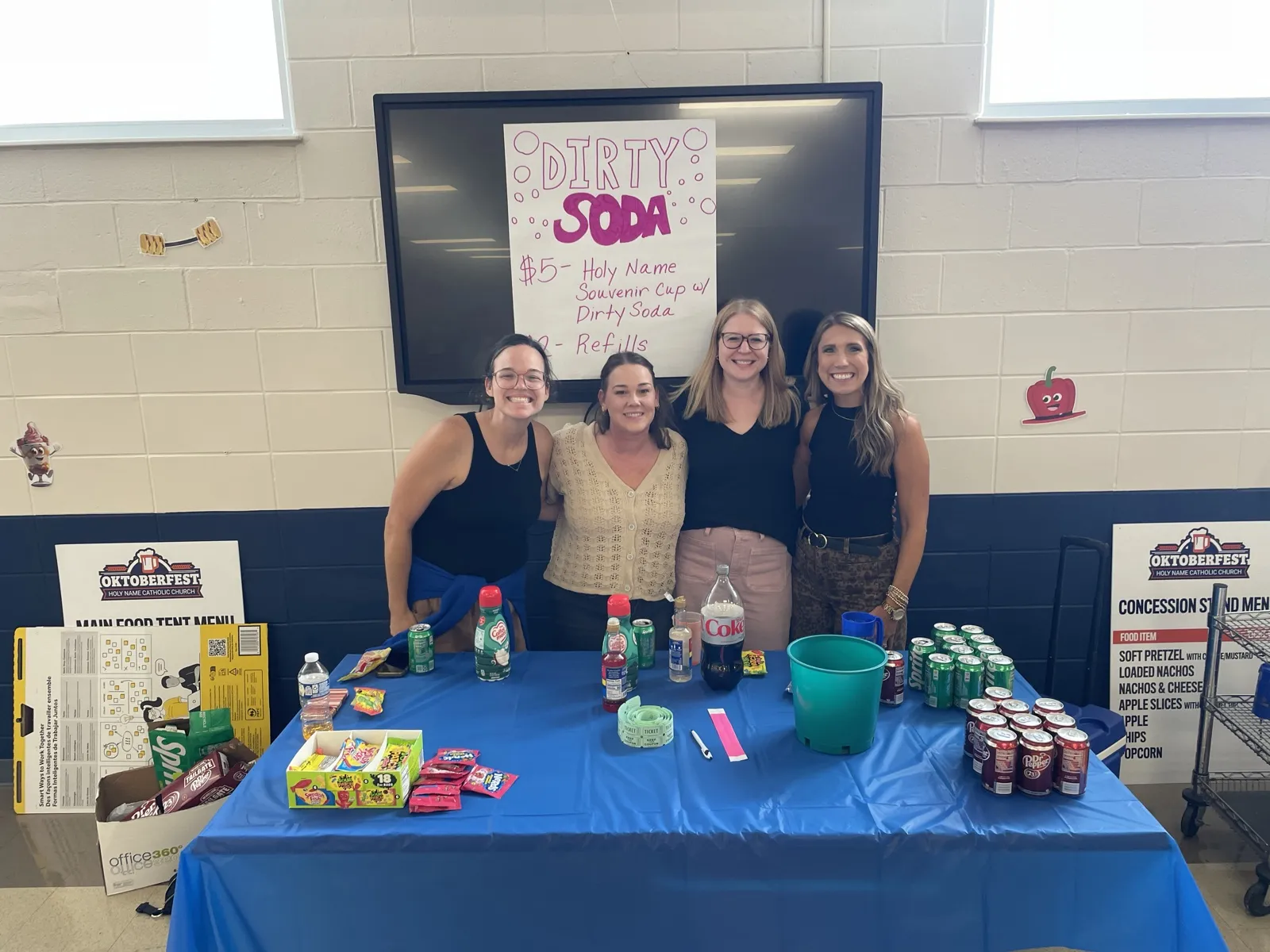 Four volunteers behind the Dirty Soda stand with a hand-lettered sign and souvenir cups
